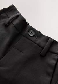 Ceinture de pantalon noire avec fermeture à bouton, boutonnière, passants de ceinture et couture visible sur fond clair.