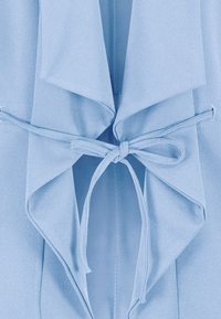 Vaaleansininen liivi, jossa on roikkuva etuosa ja solmimisnuora. Valmistettu pehmeästä kankaasta, jossa on värikäs pinta, ja siinä on kerrokselliset, virtaavat kaulukset.