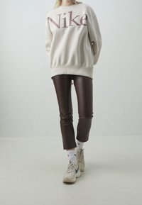 Light beige sweatshirt with large maroon "Nike" logo. Brown fitted faux leather pants. White athletic socks. Light-colored sneakers.