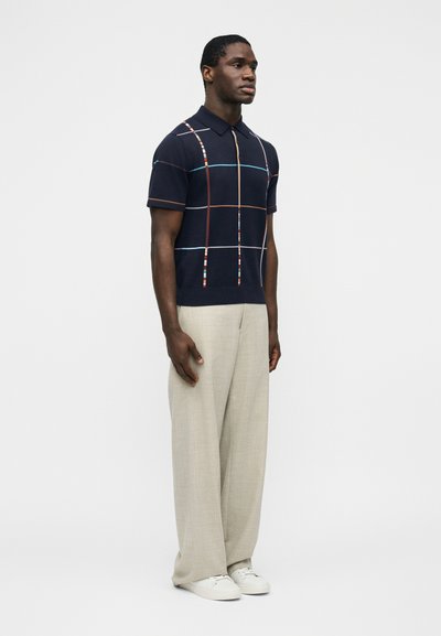 Homme debout portant un polo manches courtes bleu marine avec des rayures verticales et horizontales multicolores, un pantalon beige à jambes larges, et des baskets blanches.
