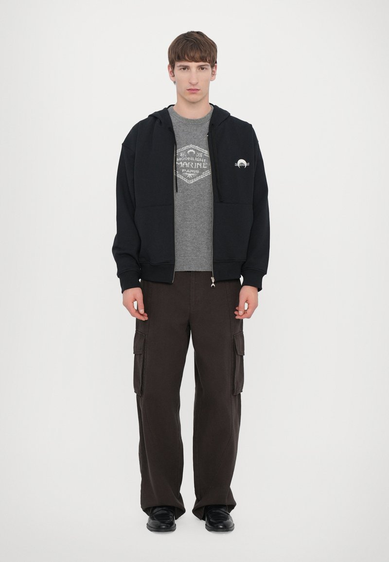 Felpa nera con zip sopra una t-shirt grafica grigia, abbinata a pantaloni cargo larghi di colore marrone scuro e scarpe nere, su sfondo bianco.
