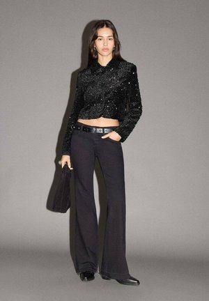 Giacca cropped nera con paillettes e colletto, abbinata a pantaloni neri a campana e una borsa coordinata; texture liscia e silhouette aderente.