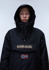 Chaqueta negra de pullover hecha de material impermeable. Cuenta con un alto cuello, un bolsillo con cremallera y un parche de la bandera noruega en la parte frontal.
