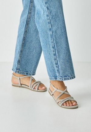 Sive sandale z trakovi z ravnim podplatom, z vzorci križanih trakov in teksturiranim poudarkom. Nošene z svetlo modrimi širokimi kavbojkami.