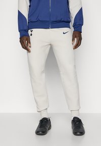 Beige joggers med elastiske manchetter, Nike-logo og broderet Tottenham Hotspur-emblem. Båret med sorte træningssko.