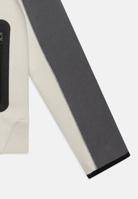 Ljusgrå och mörkgrå sweatshirt med dragkedjeficka, kontrasterande sömmar och en texturerad tygfinish.