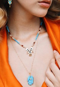 Les bijoux en or et turquoise présentent des colliers superposés avec un design géométrique, un motif de main et des accents de perles dans des couleurs variées.
