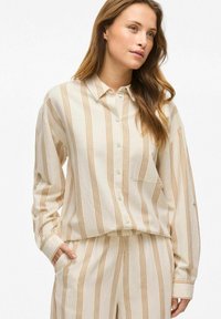 Camisa beige de botones fabricada con una tela suave, con rayas verticales naranjas y un único bolsillo en el pecho. Corte holgado con mangas con puños.
