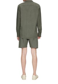 Man in olijfgroene lange mouwen shirt en bijpassende korte broek, staand met zijn rug naar de camera tegen een witte achtergrond.