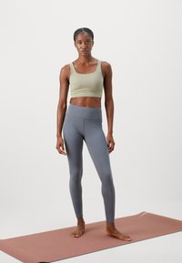 Lys grøn atletisk crop top med brede stropper, parret med højttalende grå leggings. Modellen står på en pink yogamåtte.