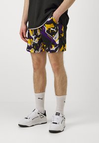 Bermudas atléticas negras con un patrón abstracto colorido en púrpura, amarillo y blanco. Incluyen bolsillos laterales y un ajuste relajado. Se combinan con zapatillas blancas.