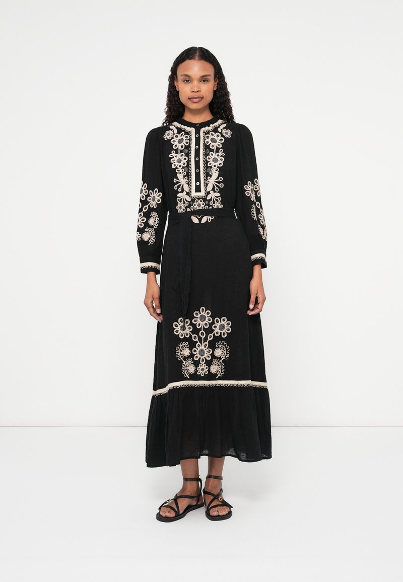 Fekete maxi ruha hosszú ujjal, fehér virágmintás hímzéssel, formált felső résszel, gombos záródással és réteges szoknyával. Fekete szandálokkal viselve.