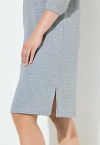 Šedé šaty s texturovanou látkou, tříčtvrtečními rukávy a postranním rozparkem. Hladké švy a přiléhavý střih zvýrazňují design.