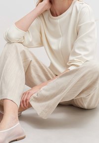 Femme assise par terre portant un haut à manches longues couleur crème, un pantalon large beige clair texturé et des chaussures blanches transparentes sans lacets.