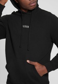 Black hoodie made of cotton with a front pocket and drawstrings. Features a "GUESS" logo in white on the chest. Soft texture, fitted design.