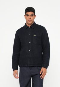 Zwart overhemdjack van een wolblend met een knoopsluiting aan de voorkant, een puntkraag en een borstzak. Heeft een klein groen logo aan de linkerkant.