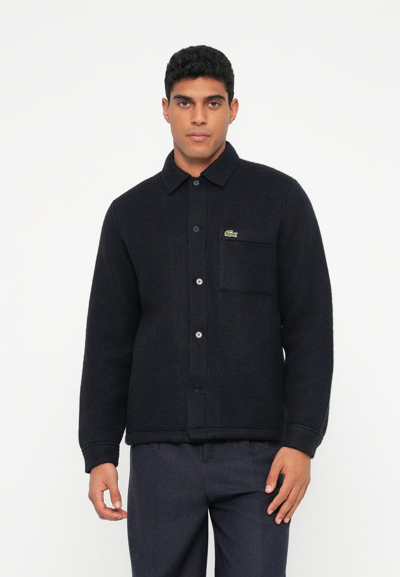 Zwart overhemdjack van een wolblend met een knoopsluiting aan de voorkant, een puntkraag en een borstzak. Heeft een klein groen logo aan de linkerkant.