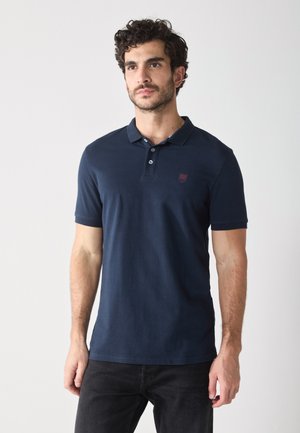 Polo shirt bleu marine en coton avec un col classique, trois boutons, des manches courtes et un petit emblème rouge sur la poitrine gauche.
