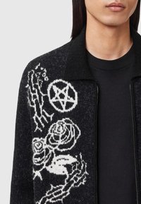 Zwarte cardigan met witte bloemen- en symboolpatronen, met een textuurafwerking en een kraaghalslijn, open gedragen over een zwart shirt.