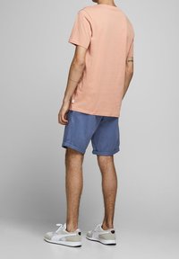 Světle růžové bavlněné tričko, krátké rukávy, v kombinaci s modrými šortkami. Model má na sobě bílé tenisky se šedými akcenty. Neformální outfit, minimalistický design.