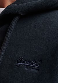 Primo piano di tessuto in pile scuro con logo SuperDry ricamato e caratteri giapponesi in filo scuro coordinato.