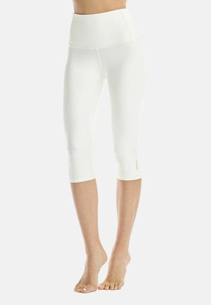 Woman wearing high-waisted, white cropped leggings standing barefoot against a plain background.