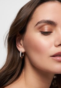 Orecchino a cerchio in oro con una fila di strass trasparenti sul davanti. Finitura metallica liscia con forma circolare. Luce che si riflette sulle pietre.