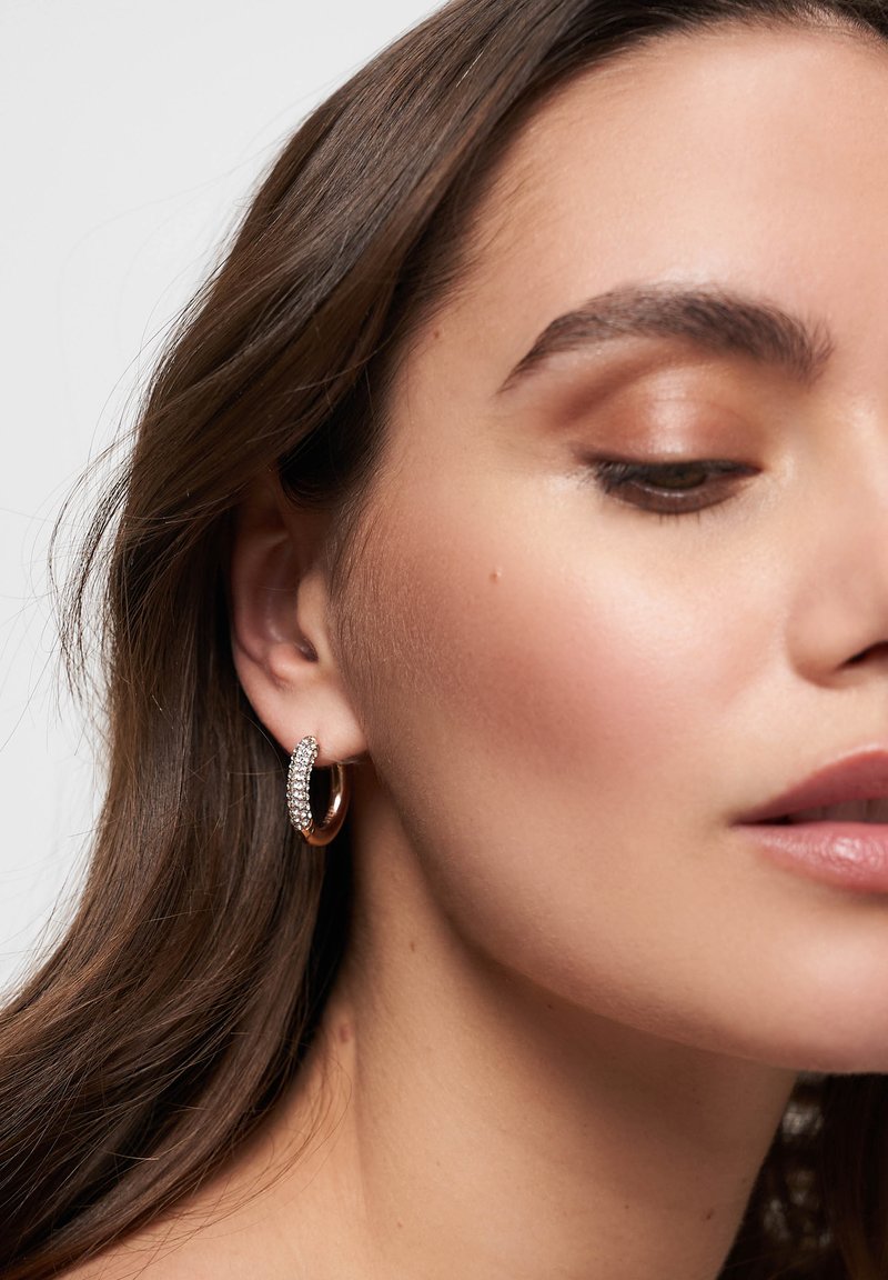 Orecchino a cerchio in oro con una fila di strass trasparenti sul davanti. Finitura metallica liscia con forma circolare. Luce che si riflette sulle pietre.