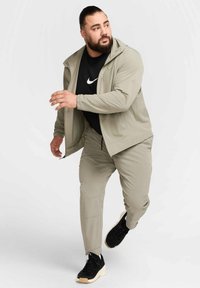 Világos olajzöld sportdzseki és nadrág, fekete Nike logós pólóval párosítva, fekete sportcipővel. Sima textúra, funkcionális dizájn.
