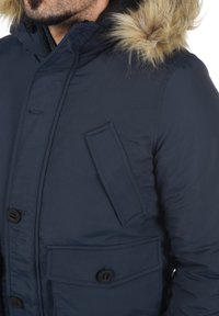 Granatowa parka z kapturem i sztucznym futrem na wykończeniach, z teksturowanym materiałem, dwoma przednimi kieszeniami oraz czarnymi guzikami do zapięcia.