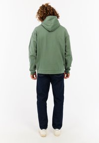 Groene katoenen hoodie met een trekkoordkap, ontspannen pasvorm, ribgebreide manchetten en onderrand. Gecombineerd met donkere denim jeans en witte sneakers.
