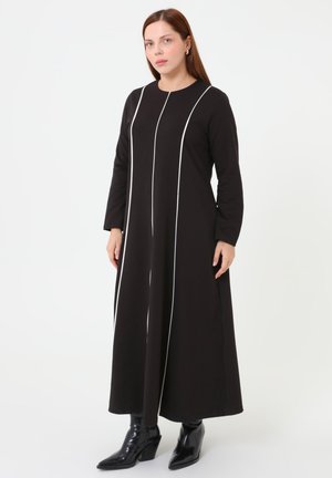 Vrouw in een lange zwarte jurk met witte verticale strepen, staand en zwarte puntige laarzen dragend tegen een effen witte achtergrond.