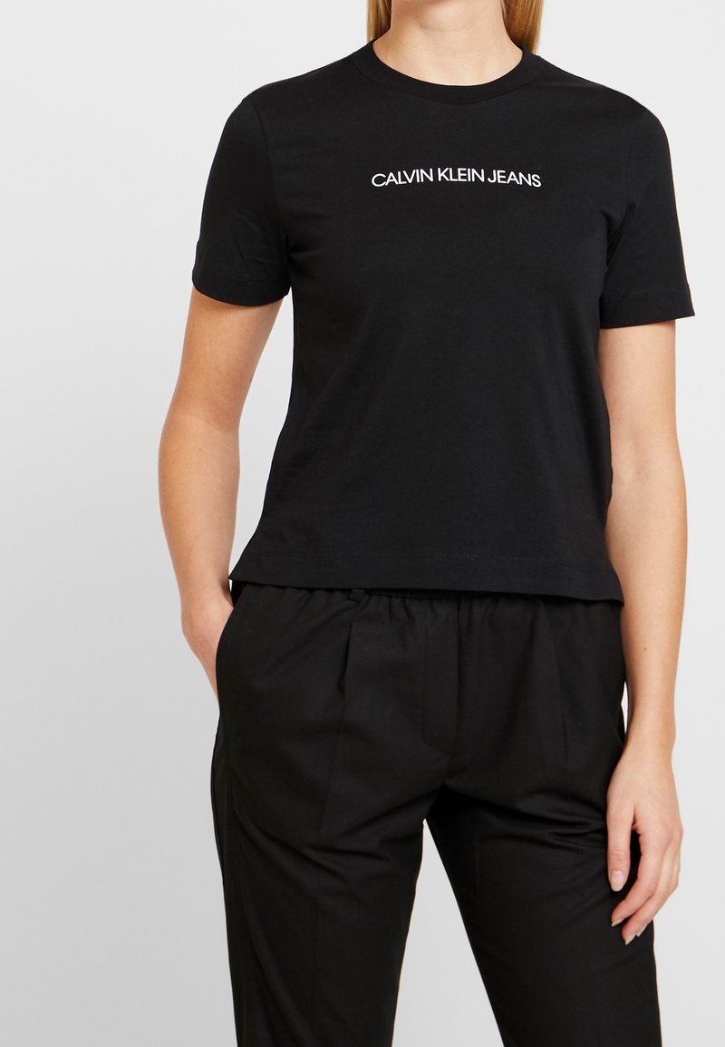 Osoba ubrana w czarną koszulkę z krótkim rękawem z napisem "CALVIN KLEIN JEANS" i czarne spodnie plisowane, pozująca z jedną ręką w kieszeni.