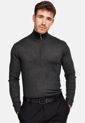Mørkegrå sweater med lynlås i strikstof, der har en tætsiddende pasform, lange ærmer og en halvlange lynlås ved kraven. Båret med sorte bukser og bælte.