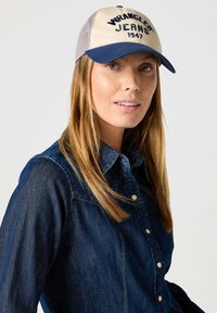 Gorra de camionero con la parte frontal beige y la visera azul marino, con bordado de "Wrangler Jeans 1947". Llevada con una camisa de mezclilla oscura.