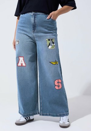 Blå jeans med vide ben og farverige bogstav-patches (A, U, S), båret med hvide og sorte sneakers og en sort top.