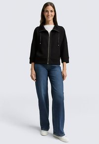 Giacca nera con cerniera e collo alto, caratterizzata da due tasche frontali e dettagli in pelle, abbinata a jeans blu a gamba larga e sneaker bianche.