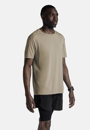 Man staat, draagt een beige kortemouwen shirt, zwarte sportshorts en een zwart polshorloge, kijkt iets naar rechts.