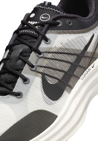 Svart och grå träningssko med en texturerad mesh-ovandel, märkets ränder och en vit sula. Skosnörena är svarta med öljetter och en logodetalj.