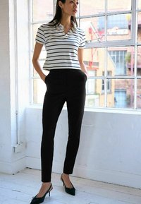 Gestreepte korte mouwen polo in wit en marineblauw, gecombineerd met een strakke zwarte broek en zwarte slingback hakken, staande bij een raam.