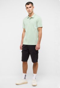 Jasnozielona koszulka polo z kołnierzykiem, zestawiona z czarnymi szortami cargo. Model nosi białe sneakersy i wzorzyste skarpetki. Prosty, casualowy styl.