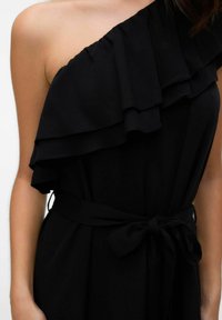 Czarna sukienka na jedno ramię z falbaniastymi detalami, cienkim paskiem w talii i gładką fakturą tkaniny. Skupienie na ramieniu i wyraźnych falbanach.