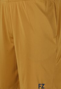 Gula träningsshorts tillverkade av ett slätt, lätt tyg. Har ett elastiskt midjeband och en liten marinblå logotyp nära fållen.
