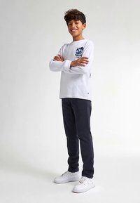 Valkoinen pitkähihainen T-paita, jossa sininen graafinen kuvio vasemmassa rinnassa, yhdistetty tummiin housuihin ja valkoisiin tennareihin.