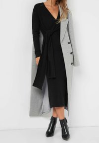 Długi szary płaszcz na czarnej sukience z wiązaniem w talii. Czarne botki do kostek dopełniają stylizację. Gładka faktura, głęboki dekolt w kształcie litery V.