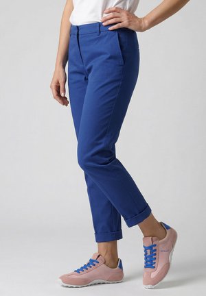 Persoon draagt opgerolde blauwe broek en roze sneakers met blauwe veters en hielaccenten, staand tegen een effen lichte achtergrond.