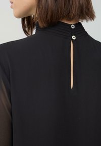 Close-up van de achterkant van de halslijn van een zwarte blouse met twee kleine knoopjes en een verticale split, gedragen door een persoon met kort bruin haar.