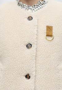 Gros plan d'une veste sherpa couleur crème avec des boutons ronds et une boucle en tissu marron avec un anneau en D doré sur la poitrine. Col de chemise à carreaux visible.