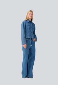 Conjunto de chaqueta de mezclilla y pantalones de pierna ancha en azul claro. La chaqueta cuenta con botones frontales y un cuello; los pantalones tienen una cintura con cordón y un ajuste holgado.
