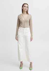 Cardigan en maille beige avec un décolleté en V profond et des boutons, associé à un pantalon large blanc et des escarpins verts à bout pointu, tissu texturé.